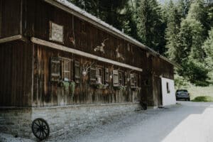 Eine rustikale Holzhütte, perfekt für einen Hochzeitsausflug, mit Geweihdekorationen und Blumenkästen unter den Fenstern inmitten eines Waldgebiets. Eine Kiesauffahrt führt zu einem in der Nähe des Gebäudes geparkten Auto. Sonnenlicht fällt durch die Bäume und wirft weiche Schatten.