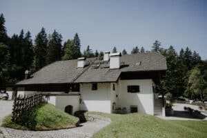 Ein rustikales Haus mit einem grauen Satteldach ist von üppigem Grün und hohen Bäumen umgeben und bietet die perfekte Kulisse für Ihre Traumhochzeit. Ein Kopfsteinpflasterweg führt unter einem klaren blauen Himmel zum Eingang und bietet eine idyllische Szene für diejenigen, die die Liebe feiern und sich auf ihre Ehereise begeben möchten.