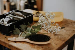 Auf einem Holztisch liegen eine Haarbürste, ein Zweig weißer Schleierkrautblüten, Eukalyptusblätter und ein schwarzer Behälter voller Friseurutensilien – perfekte Details für Ihre Hochzeitsvorbereitungen. Im unscharfen Hintergrund stehen Flaschen und eine gelbe Schale, die Ihrer Hochzeitsplanung subtile Eleganz verleihen.