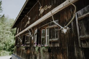 Eine rustikale Holzhütte mit Geweihen an der Außenseite und bunten Blumen in Blumenkästen bietet eine bezaubernde Kulisse für jede Hochzeit. Üppige grüne Bäume wiegen sich unter einem klaren Himmel und bilden eine perfekte Kulisse für Ihre ideale Hochzeitsplanung.