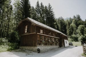 Eine rustikale Holzhütte, umgeben von hohen Kiefern, schafft eine idyllische Kulisse für die Hochzeitsplanung. Das schräge Dach und das Steinfundament der Hütte ergänzen die ruhige Atmosphäre. Ein Kiesweg führt zum Eingang, wo das Sonnenlicht durch die Bäume diesen perfekten Ort zum Feiern von Hochzeiten erhellt.