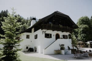 Ein charmantes, rustikales Chalet mit dunklem Holzdach und weißen Wänden ist ein idyllischer Ort für Ihre Hochzeit. Eingebettet zwischen üppigem Grün und Bäumen bietet die Terrasse Sitzgelegenheiten im Freien. Der klare, sonnige Himmel unterstreicht die friedliche Bergkulisse – perfekt für die Planung Ihrer Traumhochzeit.