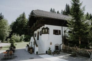 Ein rustikales zweistöckiges weißes Haus mit Holzakzenten und einem schrägen Dach, umgeben von Bäumen – ein perfekter Ort für eine bezaubernde Hochzeit. Es gibt eine Treppe, die in den zweiten Stock führt, Sitzgelegenheiten im Freien mit Tischen und im Hintergrund üppiges Grün unter einem klaren Himmel.