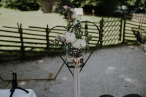 Ein herzförmiger Metallständer hält elegant eine Vase mit weißen und rosa Blumen, umgeben von üppigem Grün. Ein zartes Herzornament hängt an der Vase und verleiht ihr Charme. Vor einem rustikalen Holzzaun und sonnenbeschienenen Bäumen ist es eine perfekte Ergänzung für jede Hochzeitsplanung oder romantische Hochzeit im Freien.