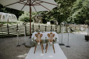 Ein bezauberndes Hochzeits-Setup im Freien besteht aus zwei Holzstühlen, die mit Blumenarrangements und Schildern mit der Aufschrift „Mr“ und „Mrs“ geschmückt sind. Ein weißer Sonnenschirm, dekorative Pflanzen, ein Holzzaun und üppiges Grün vervollständigen diese malerische Szene, perfekt für Ihre Hochzeitsplanung.