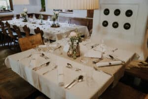 Ein wunderschön gedeckter Esstisch mit weißen Tischdecken, elegantem Glasgeschirr und Blumenschmuck schmückt einen gemütlichen Raum – perfekt für eine Hochzeit. Holzstühle umgeben den Tisch, während sanfte Beleuchtung Wärme und Charme verleiht und ein einladendes Ambiente schafft, das sich ideal für jede Hochzeitsplanung eignet.