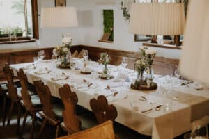 Ein langer, eleganter Esstisch ist mit weißen Tischdecken, Blumendekorationen und ordentlich angeordnetem Besteck und Gläsern gedeckt – perfekt für eine Hochzeitsplanung. Holzstühle und Zimmerpflanzen sorgen für eine gemütliche Atmosphäre, während natürliches Licht durch die nahe gelegenen Fenster strömt.