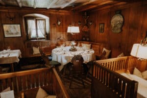 Ein gemütlicher, rustikaler Speisesaal mit Holzwänden und -decke, perfekt für eine Hochzeitsfeier. Er verfügt über runde Tische mit weißen Tischdecken, ordentlich angeordneten Stühlen und sanfter Umgebungsbeleuchtung. Dekorative Gegenstände wie Geweihe und Gemälde schmücken die Wände und schaffen eine warme Atmosphäre, die ideal für die Hochzeitsplanung ist.