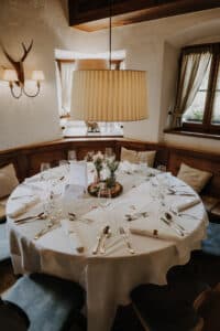 Ein runder Tisch für ein formelles Abendessen in einem gemütlichen Raum, perfekt für die Hochzeitsplanung, verfügt über eine weiße Tischdecke, ordentlich angeordnetes Besteck, Gläser, ein Menü und ein Blumengesteck. Der Raum besticht durch Holzakzente, Wanddekor und natürliches Licht vom Fenster.