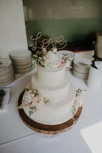 Eine zweistöckige Hochzeitstorte mit weißem Zuckerguss und rosa und weißen Blumen verkörpert perfekt die Essenz einer Hochzeit. Auf einem Metallaufsatz steht in Schreibschrift „Love“. Die Torte steht auf einem runden Holzsockel, umgeben von Stapeln von Tellern und Besteck auf einem Tisch.