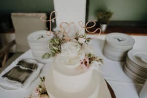 Eine zweistöckige weiße Torte, geschmückt mit weißen und rosa Blumen und einem Drahtaufsatz mit der Aufschrift „Love“, schafft die perfekte Kulisse für eine romantische Hochzeit. Sie steht auf einem Tisch mit gestapelten Tellern und Besteck im Hintergrund, ideal für Ihre sorgfältige Hochzeitsplanung.