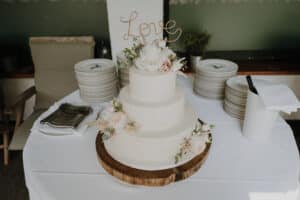Eine dreistöckige weiße Hochzeitstorte, geschmückt mit rosa und weißen Blumen, steht auf einem Holzsockel und verkörpert elegante Hochzeitsstimmung. Das Wort „Love“ ziert die Oberseite, während Teller und Servietten ordentlich dahinter auf einem weiß gedeckten Tisch angeordnet sind – perfekt für die Hochzeitsfeier eines jeden Paares.