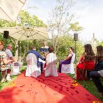 Eine Gruppe von Menschen sitzt im Freien auf hölzernen und bedeckten Stühlen, vor einem Redner - wahrscheinlich Erwin Eisenschin -, der anscheinend eine Hochzeit oder Zeremonie abhält. Rote Blütenblätter und gelbe Blumen schmücken einen roten Teppich im Gras.