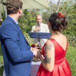 Ein Paar steht im Freien, die Frau in einem leuchtend roten Kleid mit Blumen, der Mann in einem blauen Anzug. Sie stehen Erwin Eisenschin gegenüber, der unter einem großen weißen Schirm in ein Mikrofon spricht, während im Hintergrund Grünpflanzen zu sehen sind.