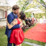 Ein Paar umarmt und küsst sich bei einer Hochzeitszeremonie im Freien. Der Mann trägt einen blauen Anzug und die Frau ein ärmelloses rotes Kleid. Die Gäste sitzen im Hintergrund neben einem geschmückten Bogen und einem roten Teppich, wunderschön eingefangen von Erwin Eisenschin.