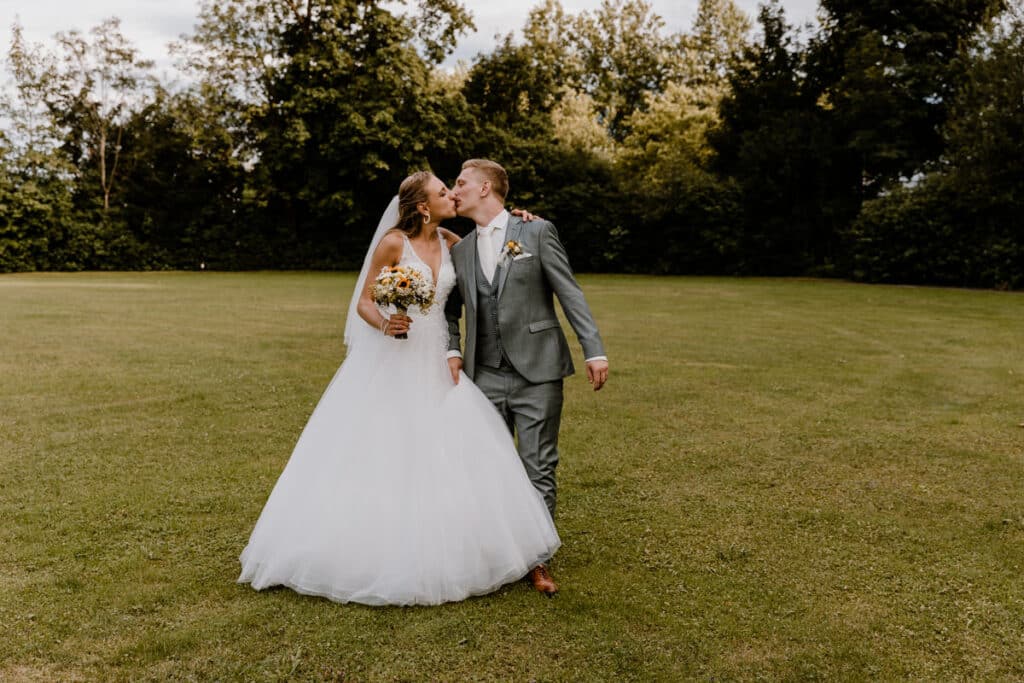 Während ihrer Hochzeit küssen sich eine Braut im weißen Kleid und ein Bräutigam im grauen Anzug, während sie Hand in Hand über einen üppigen grünen Rasen gehen. Bäume säumen den Hintergrund unter dem teilweise bewölkten Himmel und fangen die Essenz perfekter Hochzeitsplanung ein.