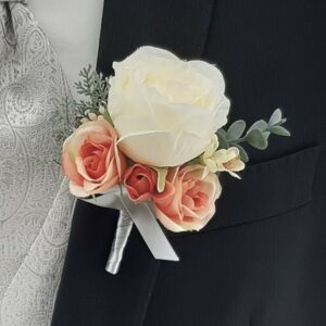 Eine Nahaufnahme einer Boutonniere, die perfekt für eine Hochzeit ist. Sie zeigt eine große weiße Rose, umgeben von kleineren pfirsichfarbenen Rosen und Grünpflanzen, mit einem silbernen Band, das anmutig um den Stiel gewickelt ist. Ideal für alle, die gerade mitten in der Hochzeitsplanung stecken.