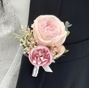 Eine Nahaufnahme des Anstecksträußchens eines Bräutigams an einem schwarzen Anzugsjackett, perfekt für die Hochzeitsplanung. Das Anstecksträußchen zeigt eine große rosa Rose, eine kleinere rosa Blume und hellgrünes Blattwerk, darunter ist eine silberne Schleife zu sehen.