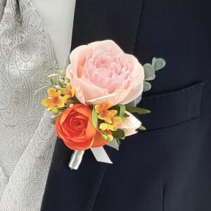 Eine Nahaufnahme eines Anzugs mit einer Boutonniere, die eine große rosa Rose, eine orange Rose, gelbe Blumen und Grünzeug zeigt, perfekt für Ihre Hochzeit. Weste und Krawatte sind silberfarben mit einem dezenten Paisleymuster.