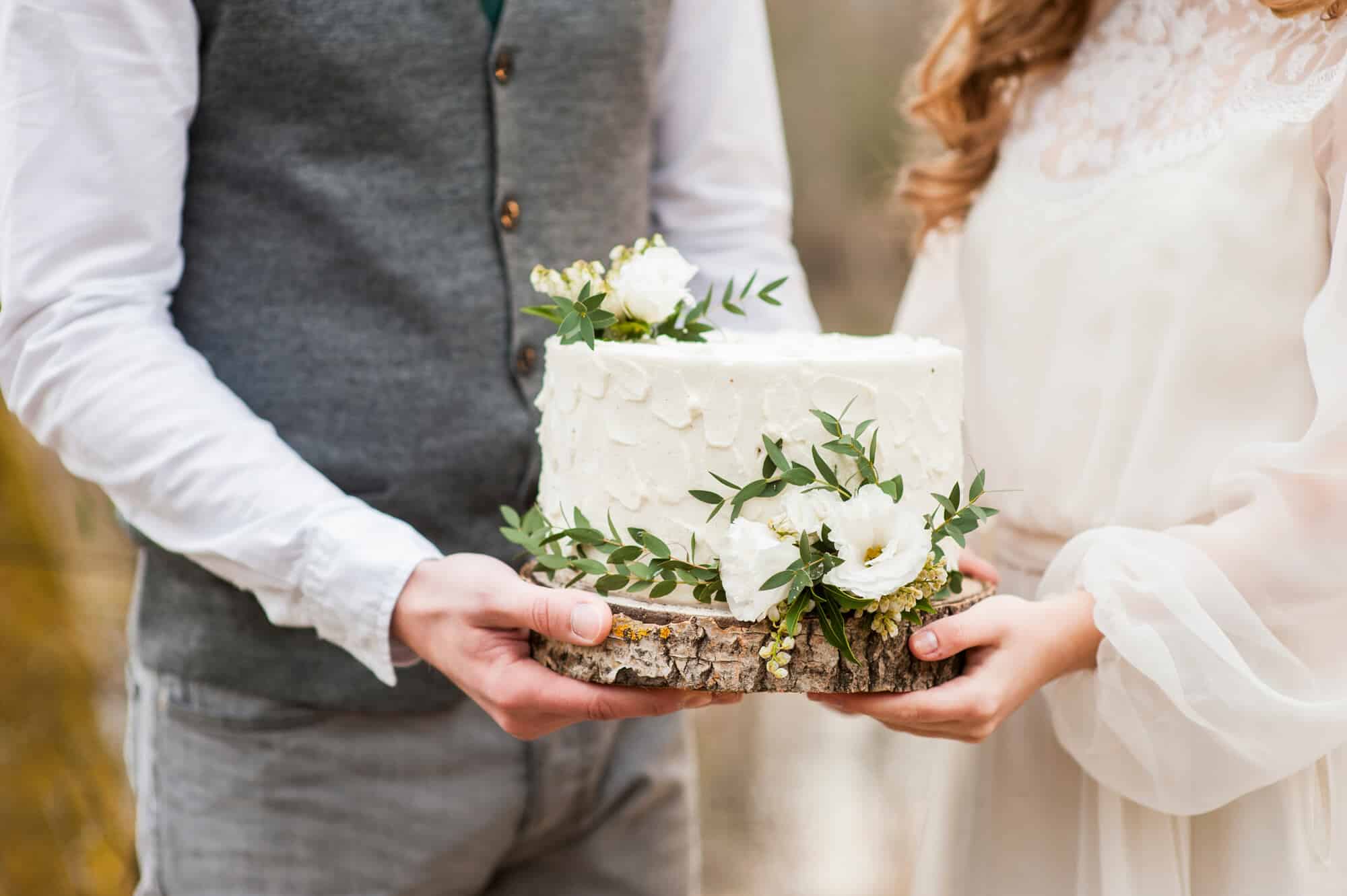 Ein Paar hält eine weiße Hochzeitstorte, die mit Grünpflanzen und kleinen weißen Blumen geschmückt ist, perfekt für ihre Hochzeit. Die Torte ruht auf einer Holzplatte. Die Person links trägt eine graue Weste, während die Person rechts ein weißes Kleid trägt.