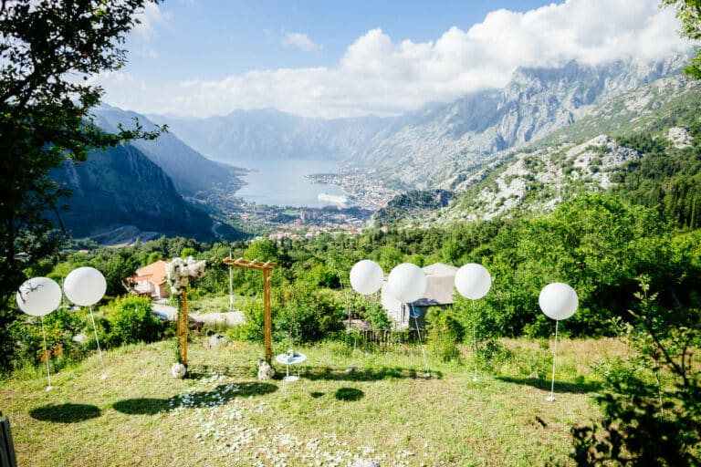 Eine malerische Hochzeitskulisse im Freien mit einem mit Blumen geschmückten Bogen, umgeben von weißen Luftballons. Perfekt für Ihre Hochzeitsplanung, bietet es eine atemberaubende Kulisse aus Bergen, einem Tal und einem See unter einem strahlend blauen Himmel – ideal für diejenigen, die planen, „Ja“ zu sagen.