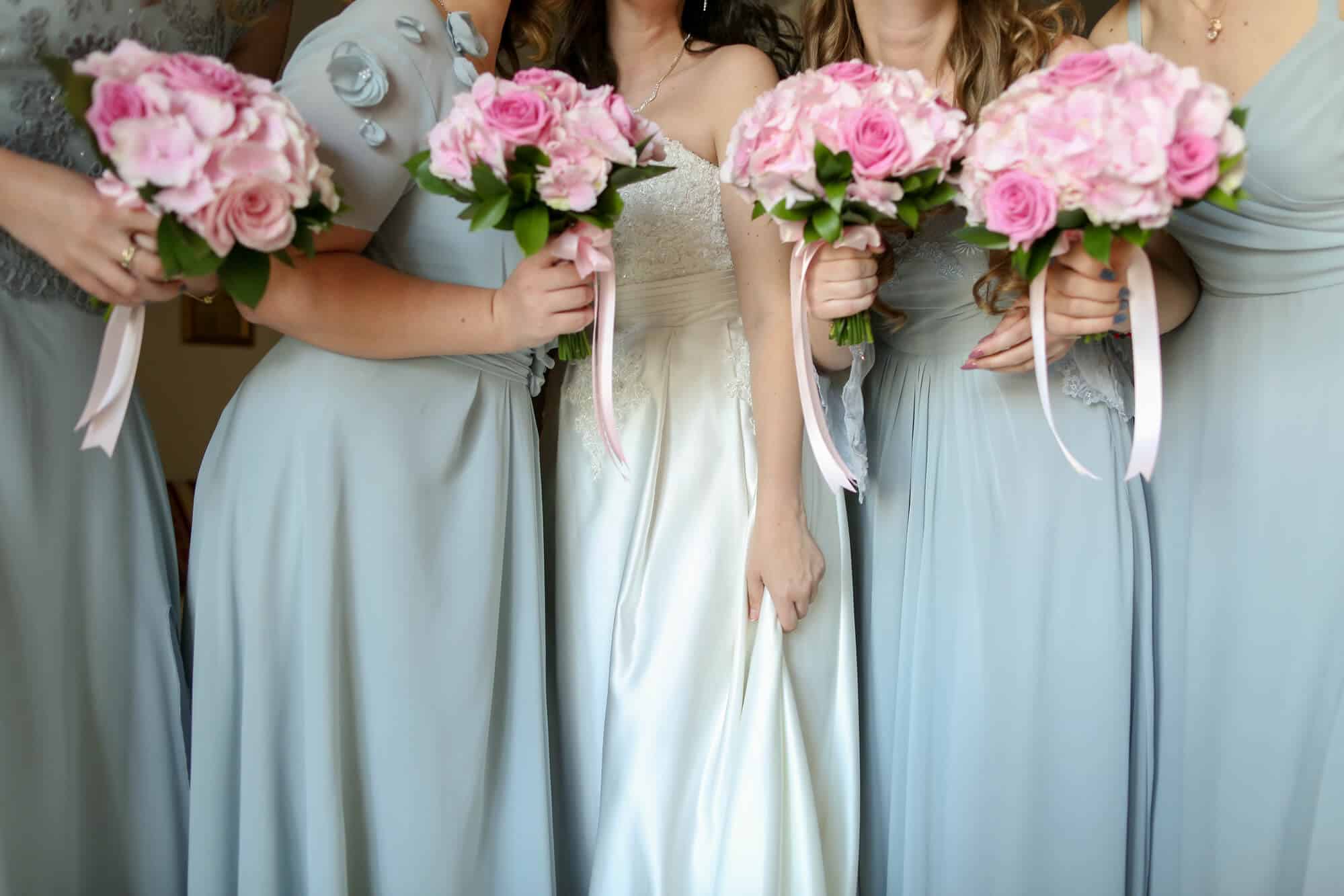 Vier Frauen in pastellfarbenen Kleidern, darunter eine Braut in Weiß, halten rosa Blumensträuße, die mit Bändern zusammengebunden sind. In einer Momentaufnahme von Eleganz und Freude posieren sie eng beieinander – ein herrlicher Moment auf der bezaubernden Reise der Hochzeitsplanung.