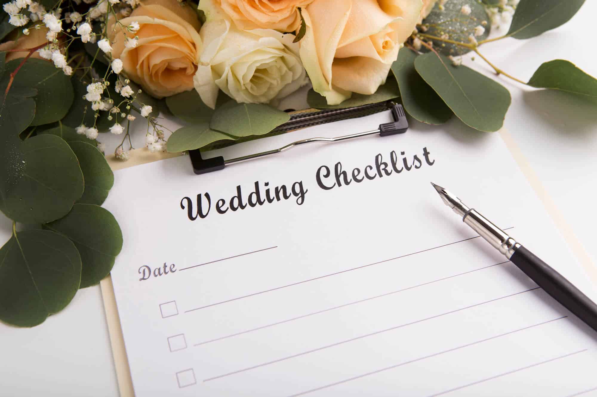 Eine Hochzeitscheckliste auf einem Klemmbrett mit leeren Zeilen und einem Stift, perfekt für Ihre Hochzeitsplanung. Das Klemmbrett ist von creme- und pfirsichfarbenen Rosen mit grünen Blättern umgeben, die eine elegante und romantische Kulisse für Ihre Hochzeitsreise schaffen.