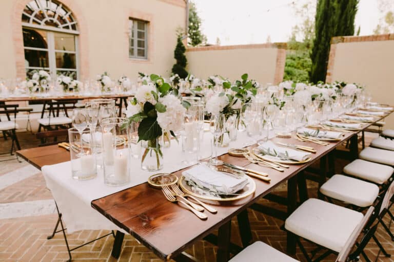 Elegantes Hochzeitsessen im Freien mit langen, in Reihen angeordneten Holztischen, perfekt für eine Hochzeitsfeier. Jeder Tisch ist mit weißen Blumendekorationen, Kerzen, Glaswaren und goldenem Besteck geschmückt. Neben den Tischen stehen ordentlich weiße Stühle mit Polstern.
