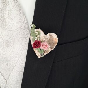 Eine Nahaufnahme einer herzförmigen Boutonniere auf einem schwarzen Anzugsjackett, perfekt für Hochzeitsfeiern. Das Herz weist eine rustikale Textur auf, die mit kleinen roten und rosa Rosen und grünen Blättern geschmückt ist und die weiße Krawatte mit Paisleymuster für einen eleganten Hochzeitsplanungslook ergänzt.