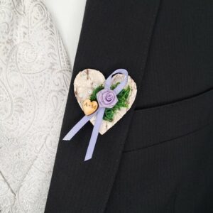 Eine herzförmige Boutonniere auf einem schwarzen Anzugjackett, perfekt für eine Hochzeit. Sie zeigt ein lila Band, eine zarte Rose, grünes Blattwerk und ein kleines Holzherz mit Initialen. Der Hintergrund ist ein weißer Stoff mit Paisleymuster, der jeder Hochzeitsplanung Eleganz verleiht.