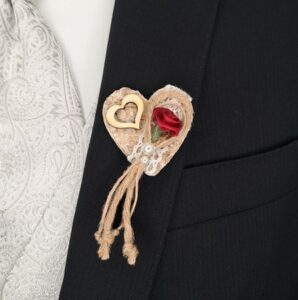 Eine Nahaufnahme eines schwarzen Anzugs, der perfekt für eine Hochzeit ist und mit einem dekorativen Herzen aus Sackleinen geschmückt ist. Dieses bezaubernde Herz besteht aus einer kleinen roten Rose, Spitze, einem hölzernen Herzakzent und Quasten aus Kordel. Perfekt kombiniert mit der hellen, gemusterten Krawatte des Anzugs für eine elegante Note bei Ihrer Hochzeitsplanung.