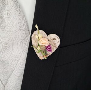 Eine Nahaufnahme eines schwarzen Anzugs mit einer herzförmigen Boutonniere am Revers, perfekt für eine Hochzeit. Die Boutonniere aus hellen Blumen und grünen Blättern ergänzt die gemusterte graue Weste elegant.
