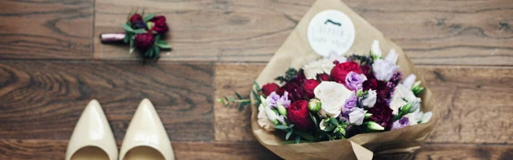 Ein Strauß roter, violetter und weißer Blumen, eingewickelt in braunes Papier, liegt auf einem Holzboden neben eleganten weißen Stöckelschuhen. Passend zu einer Traumhochzeit liegt daneben eine kleine passende Ansteckblume, die der Szene einen Hauch sorgfältiger Hochzeitsplanung verleiht.