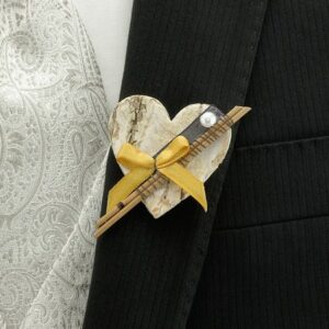 Eine Boutonniere mit herzförmigem Design und Rindenstruktur ist mit einem kleinen gelben Band und einer Perle geschmückt. Perfekt für Ihre Hochzeit, wird sie an ein schwarzes Sakko geheftet und durch eine weiße Weste mit Paisleymuster ergänzt und verleiht Ihrer Hochzeitsplanung Eleganz.