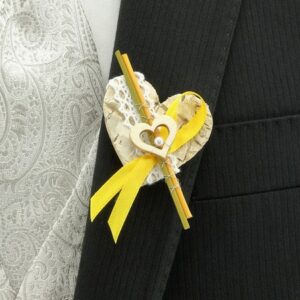 Eine Nahaufnahme eines dunklen Anzugs mit einer dekorativen herzförmigen Boutonniere, perfekt für eine Hochzeit. Die Boutonniere ist mit einem gelben Band, Spitze und einer kleinen hölzernen Herzverzierung verziert. Im Hintergrund ist teilweise eine Weste mit Paisleymuster zu sehen.