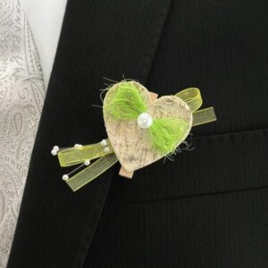 Eine herzförmige Boutonniere mit grünem Band und einer Perle ziert ein schwarzes Jackett, ergänzt durch eine verzierte Weste mit silbernem Muster darunter – perfekt für den eleganten Anlass einer Hochzeit.