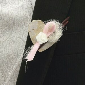 Eine Nahaufnahme eines mit einem dekorativen Boutonniere geschmückten Anzugsjacketts, perfekt für eine Hochzeit. Es verfügt über eine herzförmige Basis, eine weiße Rose, ein rosa Band und dekorative Fasern, die alle den dunklen Anzug mit einer weißen Weste mit Paisleymuster elegant ergänzen.