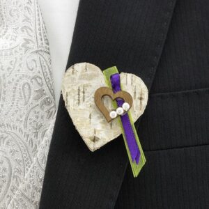 Eine Nahaufnahme eines Anzugs mit einer gemusterten hellen Krawatte, ideal für eine Hochzeit. Am Revers ist eine herzförmige Boutonniere aus Holz und Rinde befestigt, mit einem kleineren Holzherz, lila und grünem Band und kleinen weißen Perlen. Perfekt für eine durchdachte Hochzeitsplanung.