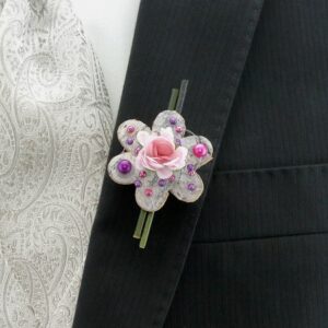 Eine Nahaufnahme eines Anzugs mit einer Krawatte mit Paisleymuster und einer Boutonniere, perfekt für eine Hochzeit. Die Boutonniere ist in Form einer Blume mit einer rosa Rose in der Mitte gestaltet, mit rosa und lila Perlen geschmückt und am Revers des dunklen Anzugs befestigt.