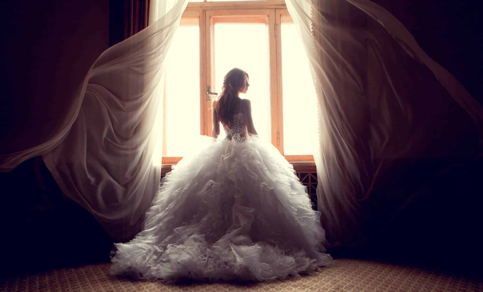 Eine Frau in einem fließenden weißen Kleid steht vor einem hellen Fenster, um das sich transparente Vorhänge bauschen, und verkörpert die Essenz einer eleganten Hochzeit. Der schwach beleuchtete Raum hebt die komplizierten Details des Kleides hervor und schafft eine heitere, ätherische Atmosphäre, die perfekt für eine durchdachte Hochzeitsplanung ist.