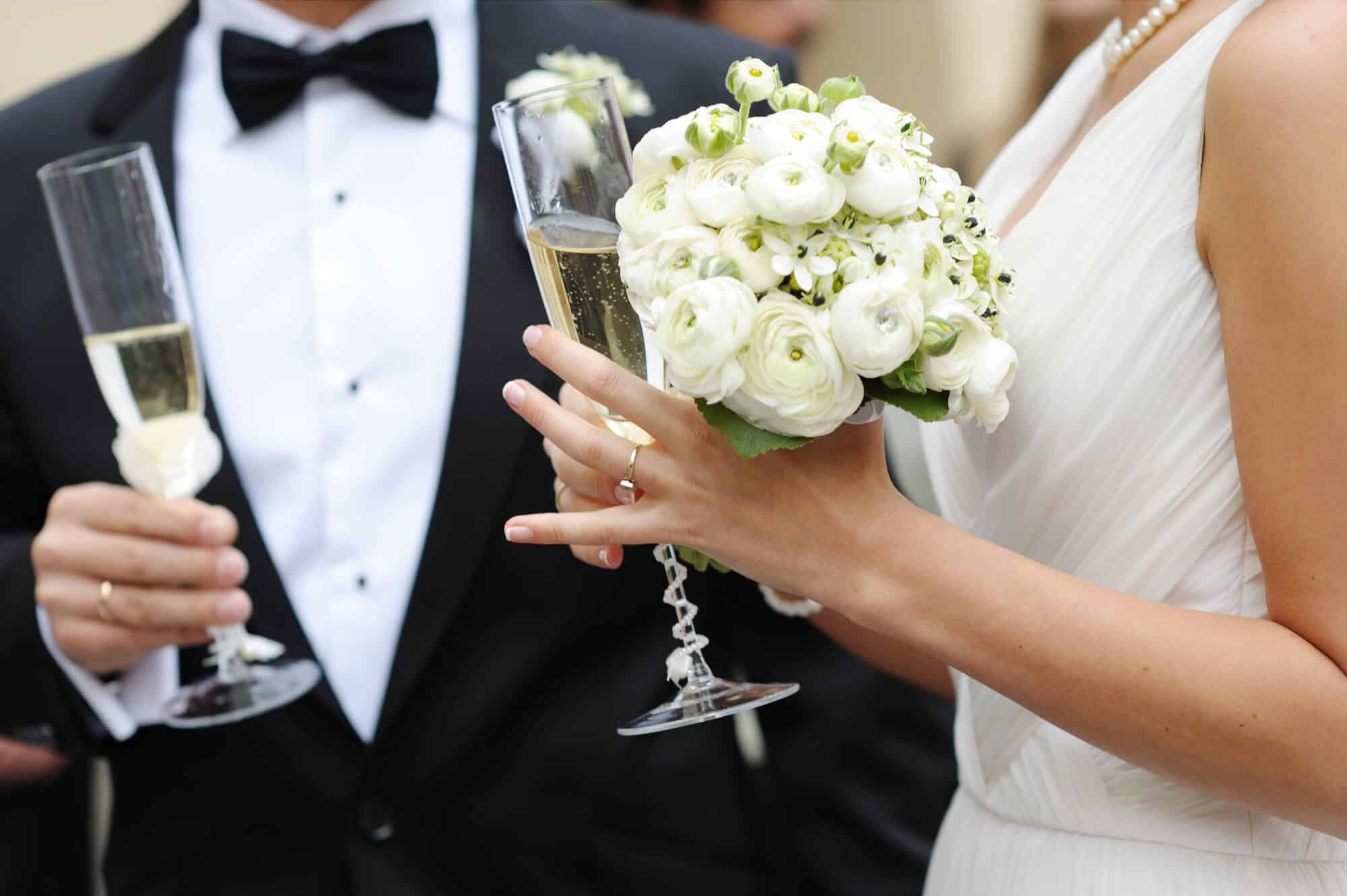Bei ihrer Hochzeit stoßen Braut und Bräutigam mit Champagnergläsern an. Die Braut, in einem weißen Kleid mit Perlenkette, hält einen Strauß weißer Blumen, während der Bräutigam im schwarzen Smoking neben ihr steht.