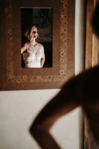 Eine Frau in einem weißen Kleid spiegelt sich in einem dekorativen Spiegel an der Wand und fängt ihre Eleganz bei der Planung ihrer Hochzeit ein. Das Bild zeigt sie von der Taille aufwärts, wobei sanftes Licht das aufwendige Design ihres Kleides hervorhebt.