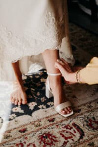 Zur Vorbereitung auf die Hochzeit befestigt eine Person in einem weißen Kleid mit Spitzenbesatz vorsichtig den Knöchelriemen einer weißen Sandale mit hohem Absatz am Fuß einer anderen Person. Dieser intime Moment spielt sich auf einem kunstvoll gemusterten Teppich ab.