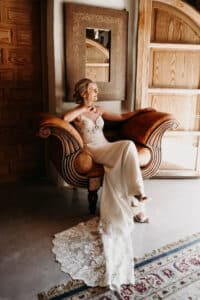 Eine Frau in einem weißen Spitzenkleid sitzt auf einem verzierten Holzstuhl und denkt über ihre bevorstehende Hochzeit nach. Der Raum mit seinen Holztüren und dem gemusterten Teppich ist in Sonnenlicht getaucht. Ihre Haare sind hochgesteckt und fangen die Essenz ihres besonderen Tages ein, während sie aus dem Fenster blickt.