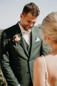 Ein lächelnder Bräutigam im grünen Anzug, geschmückt mit einer Boutonnière und einer Fliege, steht der Braut gegenüber, die teilweise von hinten zu sehen ist. Es ist ein perfekter sonniger Tag für ihre Hochzeit, da sie ihre Liebe im Freien feiern.