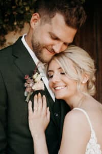 Ein Paar umarmt sich liebevoll und ist in die Freude seiner Hochzeit versunken. Die Frau im weißen Kleid lehnt ihren Kopf an die Brust des Mannes und lächelt mit geschlossenen Augen. Der Mann im grünen Anzug lächelt herzlich. An seinem Anzug ist eine Ansteckblume mit Blumen befestigt, während sie von Heiratsträumen umgeben sind.