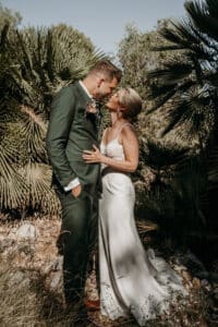 Inmitten üppigen Grüns stehen Braut und Bräutigam kurz davor, sich zu küssen. Dies ist ein intimer Moment ihrer Hochzeit. Der Bräutigam trägt einen grünen Anzug, während die Braut ihr weißes Kleid trägt. Sie verkörpern Romantik und Eleganz, während sie ihre Liebe feiern.