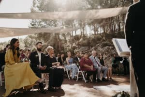 Eine Hochzeitszeremonie im Freien unter dreieckigen Schirmen, durch die das Sonnenlicht fällt. Die Gäste sitzen auf weißen Stühlen und blicken aufmerksam auf den Geistlichen. Bäume und Hügel bilden den Hintergrund und schaffen eine malerische, rustikale Atmosphäre, ideal für diejenigen, die sich leidenschaftlich mit Hochzeitsplanung beschäftigen.