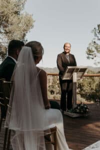 Braut und Bräutigam sitzen bei einer Hochzeit im Freien einem lächelnden Geistlichen gegenüber. Die Braut trägt einen Schleier und ein weißes Kleid, der Bräutigam einen dunklen Anzug. Sie stehen auf einer Holzplattform, umgeben von Bäumen unter einem klaren Himmel, und verkörpern ihre perfekte Hochzeitsplanung.