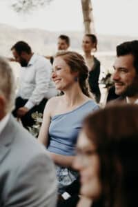 Eine Frau in einem blauen Kleid sitzt lächelnd in einer Gruppe im Freien und feiert eine Hochzeit. Die anderen um sie herum tragen formelle Kleidung und nehmen an dem freudigen Anlass inmitten von Bäumen und einer sonnigen, natürlichen Umgebung teil.
